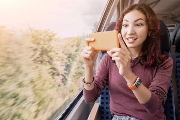 Asian girl takes photos and shoots videos on her smartphone through the window of a high-speed train during a trip or a tour