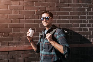 Elinde kahve kupası olan şık bir genç adam fabrikadaki turuncu tuğla duvarın arkasında poz veriyor.