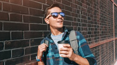 Elinde kahve kupası olan şık bir genç adam fabrikadaki turuncu tuğla duvarın arkasında poz veriyor.