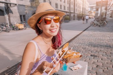 Şehir caddesinde yemek çubuklarıyla paket kutusundan börek yiyen neşeli kız. Fast food ve Asya mutfağı.