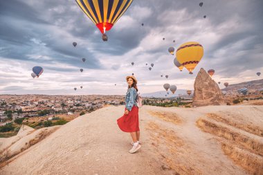 Elbisesinde güneş ışığı parıldıyor, mutlu genç kadın Cappadocia kırsalında duruyor, sıcak hava balonlarının siluetleri gökyüzünde dans ediyor..