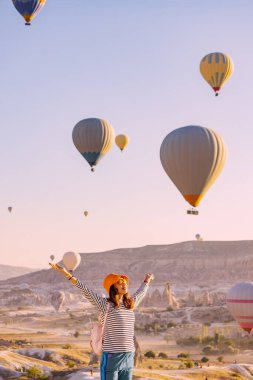 Mutlu gezgin kız el kaldırıyor ve ünlü turistik merkezde uçan sıcak hava balonları seyrediyor.