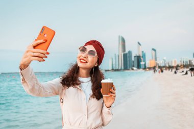 Su berrakken ve arka planda Abu Dabi 'nin gökdelenleri yükselirken, genç bir kız sahildeki akıllı telefonunda bir selfie çekiyor, anı ve çarpıcı manzarayı yakalıyor..