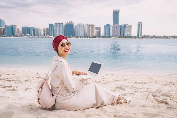 stock image With a laptop open, a young asian woman works remotely on the beach, taking advantage of the stunning view of Abu Dhabi's skyscrapers as her backdrop.