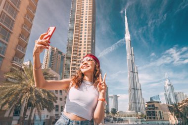Mutlu Asyalı turist kız, seyahat blogu için Dubai şehir merkezinde dünyanın en büyük gökdeleni Burj Khalifa 'nın arka planında selfie çekiyor.