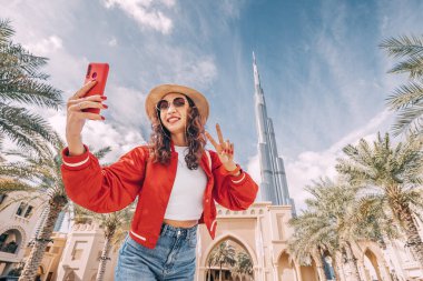 Mutlu Asyalı turist kız, seyahat blogu için Dubai şehir merkezinde dünyanın en büyük gökdeleni Burj Khalifa 'nın arka planında selfie çekiyor.