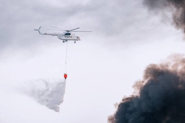 İtfaiyeci acil durum helikopteri yangını söndürüyor ve sepetteki siyah dumanı şehrin üzerine püskürtüyor.