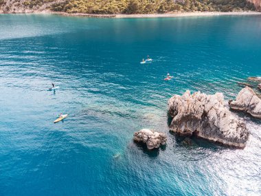Kürekli bir grup veya takımın havadan görüntüsü, deniz kenarındaki kayaların yanındaki SUP tahtalarında yüzüyor. Sağlıklı yaşam tarzı ve eğlence kavramı