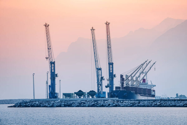 Working construction and industrial cranes at Antalya city cargo sea port with ships against mountains background at sunset. Marine shipment industry concept