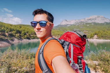 Sırt çantalı mutlu yürüyüşçü arka planda gölle dağlarda kendi fotoğrafını çekiyor.