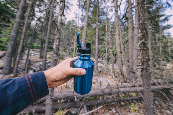 Modern metal tourist flask for drinking water against the background of a blurred forest in hand of a male hiker