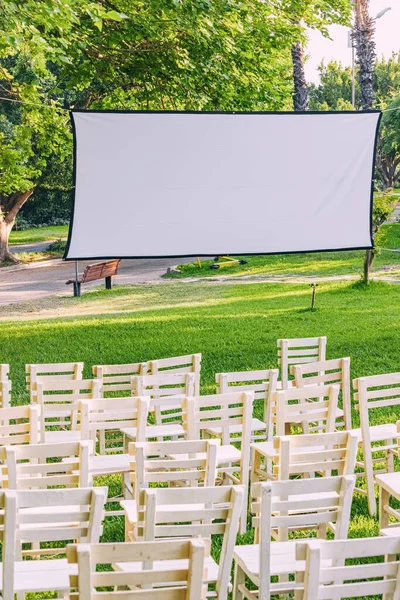 Open-Air Cinema: An empty white screen awaits projection in a park, promising summer outdoor entertainment and movie magic.