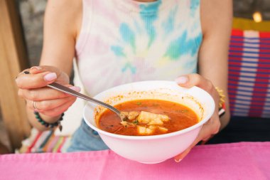 Bir kadın beyaz bir kaseden çorba içiyor. Elinde bir kaşık tutuyor.