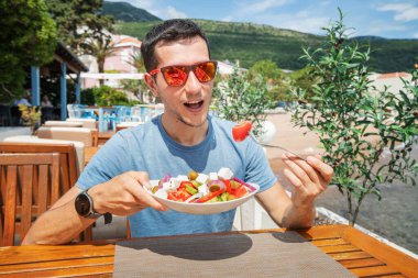 Açık havada taze Yunan salatasının tadını çıkaran bir adam. İçinde zeytin, soğan ve peynir gibi organik malzemeler var..