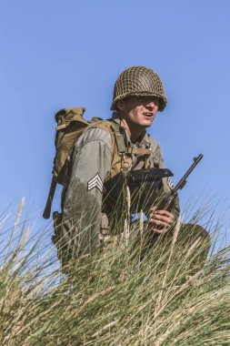 Historical reeneactor dressed as  World War II-era infantry soldier views the site while kneeling among the tall grass during a historical reenactment . Hel, Poland clipart