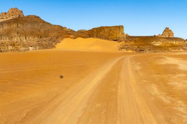 Tekerlek izleri olan kumlu toprak yol. Çöl manzarası. Gün batımında inanılmaz kum taşı oluşumları. Tadrart Rouge, Tassili N 'Ajjer Ulusal Parkı, Cezayir, Afrika.  
