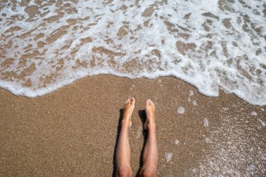Deniz dalgalı kumlu bir sahilde çıplak ayaklar. İnsanlar tatilde dinleniyor, sahilde vakit geçiriyor, yaz tatili konsepti..