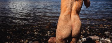 A young naked woman covered by sand walks into the sea. View from the back beauty girl outdoors enjoying nature. 