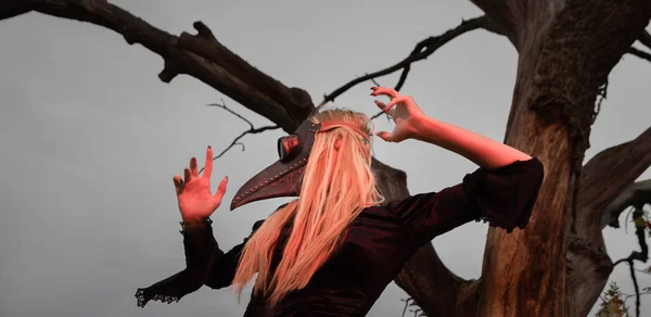 Natures beauty with a touch of mystery. A mystical and captivating image - a blonde woman in a crow mask posing near a withered tree in nature, creating an aura of mystery and beauty.