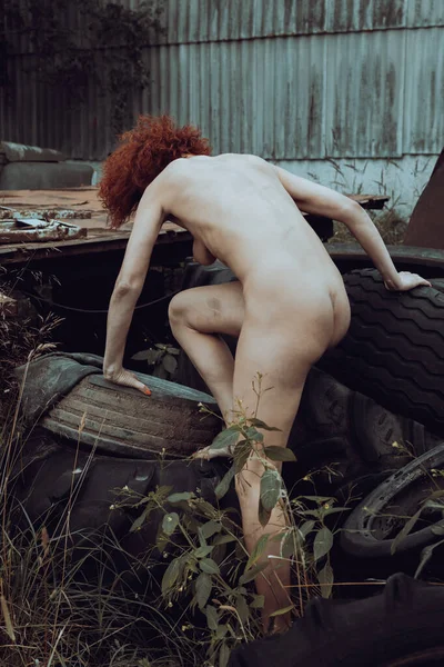 stock image A completely naked red-haired woman poses among old tires in a scrap yard and used agricultural machinery