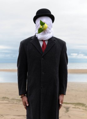 Yüzü kumaşla kaplı, şapka ve ceket giyen bir adam, deniz kıyısında ellerini kullanmadan ağzında bir elma tutuyor..