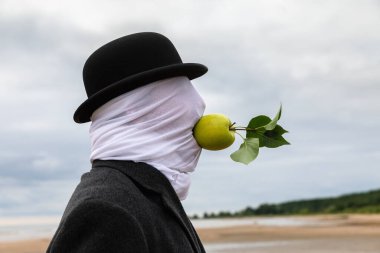 Yüzü kumaşla kaplı, şapka ve ceket giyen bir adam, deniz kıyısında ellerini kullanmadan ağzında bir elma tutuyor..