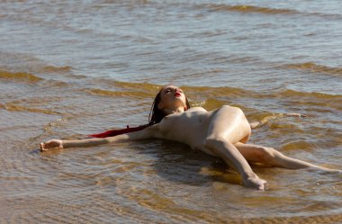 Kırmızılı, çırılçıplak genç bir kadın deniz kenarında doğanın tadını çıkarıyor.