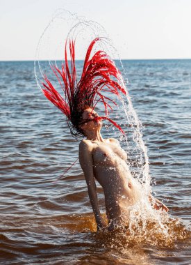 Kırmızılı, çırılçıplak genç bir kadın deniz kenarında doğanın tadını çıkarıyor.