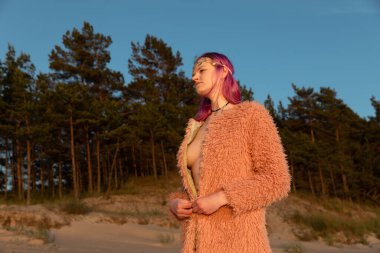 Deniz kıyısında çıplak bedenle giyinmiş genç bir kadının portresi batan güneşin ışınlarında bir çam ormanının arka planına karşı.