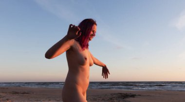 Genç ve güzel bir kadın, batan güneşin ışığında, deniz kıyısında çıplak yürüyor.