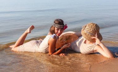 Geçmişe dönük mayolu iki genç kadın, şapka ve yelpaze, yaz günlerinde sakin bir denizin kıyısında doğanın ve birbirlerinin tadını çıkarıyorlar.