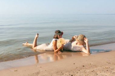 Geçmişe dönük mayolu iki genç kadın, şapka ve yelpaze, yaz günlerinde sakin bir denizin kıyısında doğanın ve birbirlerinin tadını çıkarıyorlar.