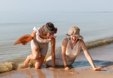 Geçmişe dönük mayolu iki genç kadın, şapka ve yelpaze, yaz günlerinde sakin bir denizin kıyısında doğanın ve birbirlerinin tadını çıkarıyorlar.