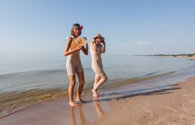 Geçmişe dönük mayolu iki genç kadın, şapka ve yelpaze, yaz günlerinde sakin bir denizin kıyısında doğanın ve birbirlerinin tadını çıkarıyorlar.