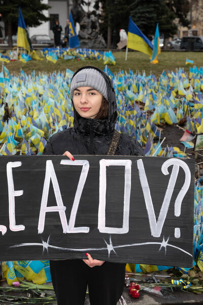 KYIV, UKRAINE - Feb. 18, 2024: A young woman is seen holding a FREE AZOV poster against the background of a large number of Ukrainian flags with the names of prisoners and the dead