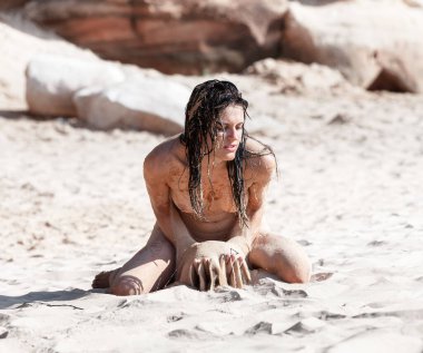 Kumsalda çıplak poz veren genç bir kadın kumla oynuyor ve doğanın tadını çıkarıyor..