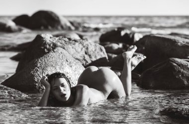 Genç ve güzel bir kadın deniz kıyısındaki büyük kayaların arasında çıplak poz veriyor. Gençlik ve çıplaklık kavramı.