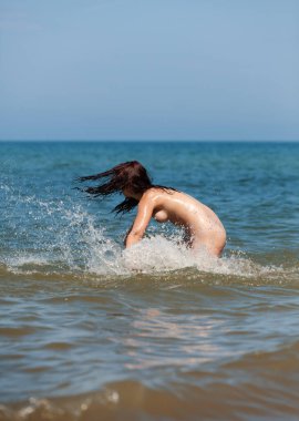 Tamamen çıplak genç bir kadın, doğa ve denizin tadını çıkarıyor, sığ sularda yürüyor ve sıçrıyor. Denizde eğlenen mutlu kadın.