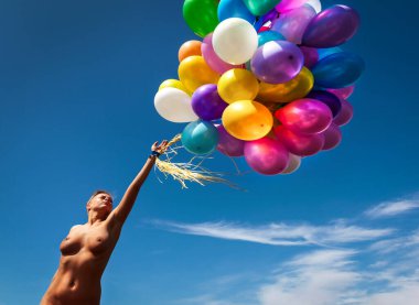 Genç bir kadın mavi gökyüzünün arka planında deniz kenarında çok renkli balonlarla çıplak poz veriyor.