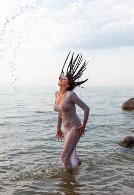 Harika vücutlu, orta yaşlı bir kadın sahilde yarı saydam bir elbiseyle poz veriyor.