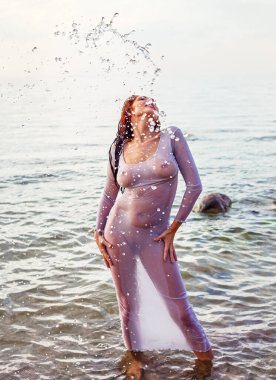 Harika vücutlu, orta yaşlı bir kadın sahilde yarı saydam bir elbiseyle poz veriyor.