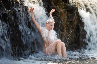 Genç ve güzel bir kadın şelalede çıplak poz veriyor ve temiz suyun tazeliğinin ve spreyinin tadını çıkarıyor.. 