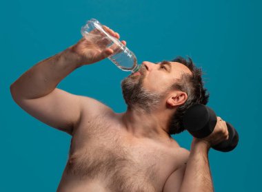 An elderly man lifts a dumbbell while drinking water to stay hydrated. He is focused on his fitness routine in a bright setting, showcasing an active lifestyle and dedication to health. clipart