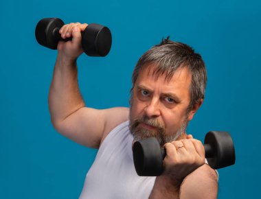 Yaşlı bir adam gücünü ve zindeliğini arttırmak için halterlerle egzersiz yapar. Onun odaklanmış ifadesi parlak renkli bir spor salonunda çalışma sırasında kararlılığını yansıtır..