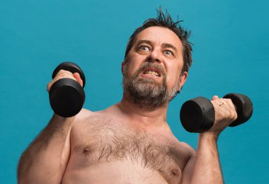 Yaşlı bir adam ağırlık kaldırırken yoğun bir şekilde odaklanarak halterlerle egzersiz yapar. Parlak mavi arkaplan, kararlı ifadesiyle çelişiyor. Gücünü ve uygunluğunu gösteriyor..