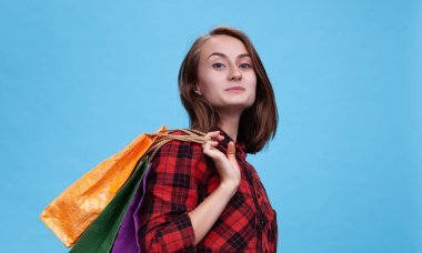 Neşeli genç bir kadın, canlı alışveriş torbalarını sergileyerek mavi bir arkaplanda kendine güvenerek duruyor. Heyecan verici bir güne hazır, ticaret sezonunun neşesini somutlaştırıyor..