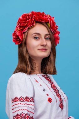 Bir kız, geleneksel Ukrayna işlemeli gömleğini sergileyerek mavi bir arka fona karşı güvenle duruyor. Çiçekli başlığı görünüşüne renk ve kültür katıyor..