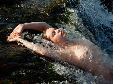 Çıplak kadın şelalede yatıyor, güneşin ve serin suyun tadını çıkarıyor..