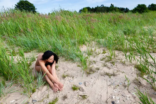 Çıplak bir kadın, uzun bitkilerle çevrili çimenli bir alanda rahatça uzanır. Sıcak güneş ışığı, sakin manzarayı yıkıyor ve gün ortasında sakin bir atmosfer yaratıyor..