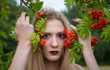 Eşsiz gözleri ve uzun saçları olan genç bir kadın elinde yeşil dallar tutarken canlı kırmızı böğürtlenler arasında poz veriyor. Atmosfer sakin, doğa ve güzelliğe odaklı..